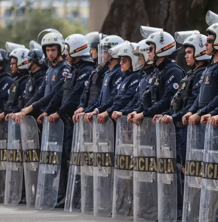 Fotografia de um pelotão de policiais militares fardados com uniformes azul-escuros e capacetes brancos. Eles estão posicionados em linha, segurando grandes escudos táticos transparentes com a inscrição "POLÍCIA" na parte inferior, formando uma barreira.