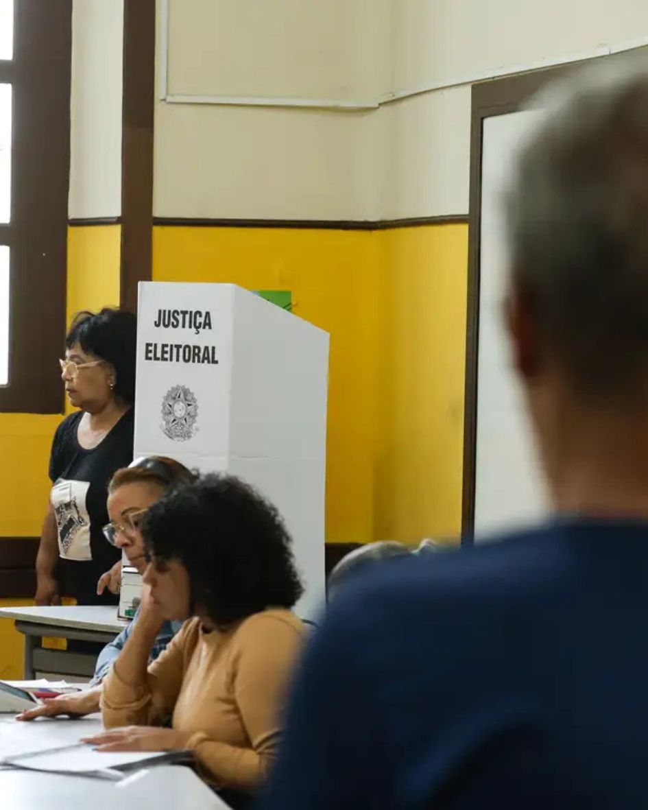 Visão do interior de uma seção eleitoral brasileira com paredes amarelas e beges. No fundo, há uma cabine de votação branca com a inscrição "JUSTIÇA ELEITORAL" e o Brasão da República. Duas mesárias estão sentadas em uma mesa com documentos em primeiro plano; uma à esquerda usa óculos e blusa listrada. À direita, um eleitor de costas está desfocado em primeiro plano, aguardando.