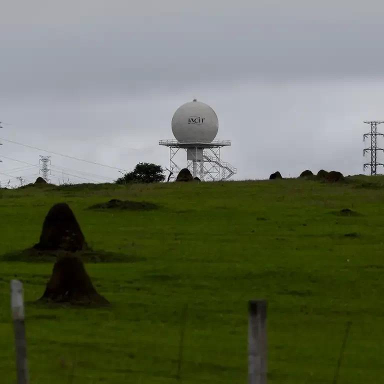 Radar meteorológico branco em formato esférico sobre uma torre metálica, localizado em um campo verde com cupinzeiros e torres de transmissão ao fundo sob céu nublado.