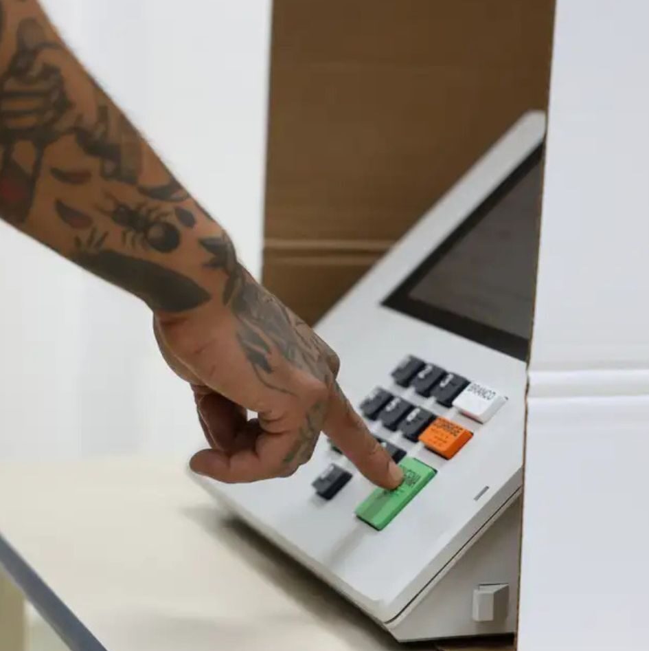 Close-up da mão direita de um eleitor com tatuagens detalhadas no antebraço, incluindo uma formiga e padrões geométricos, pressionando o dedo indicador no botão verde "CONFIRMA" de uma urna eletrônica brasileira branca. O teclado numérico e os botões coloridos "BRANCO" e "CORRIGE" são visíveis. A urna está dentro de uma cabine de votação de papelão.