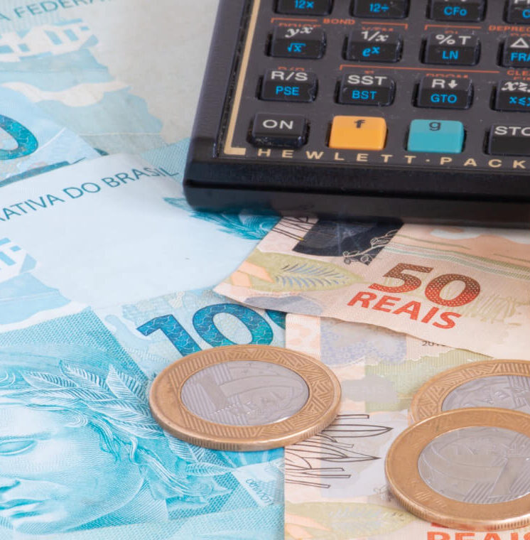 Fotografia em close-up de uma calculadora financeira preta apoiada sobre notas de cem e cinquenta Reais e algumas moedas de um Real.