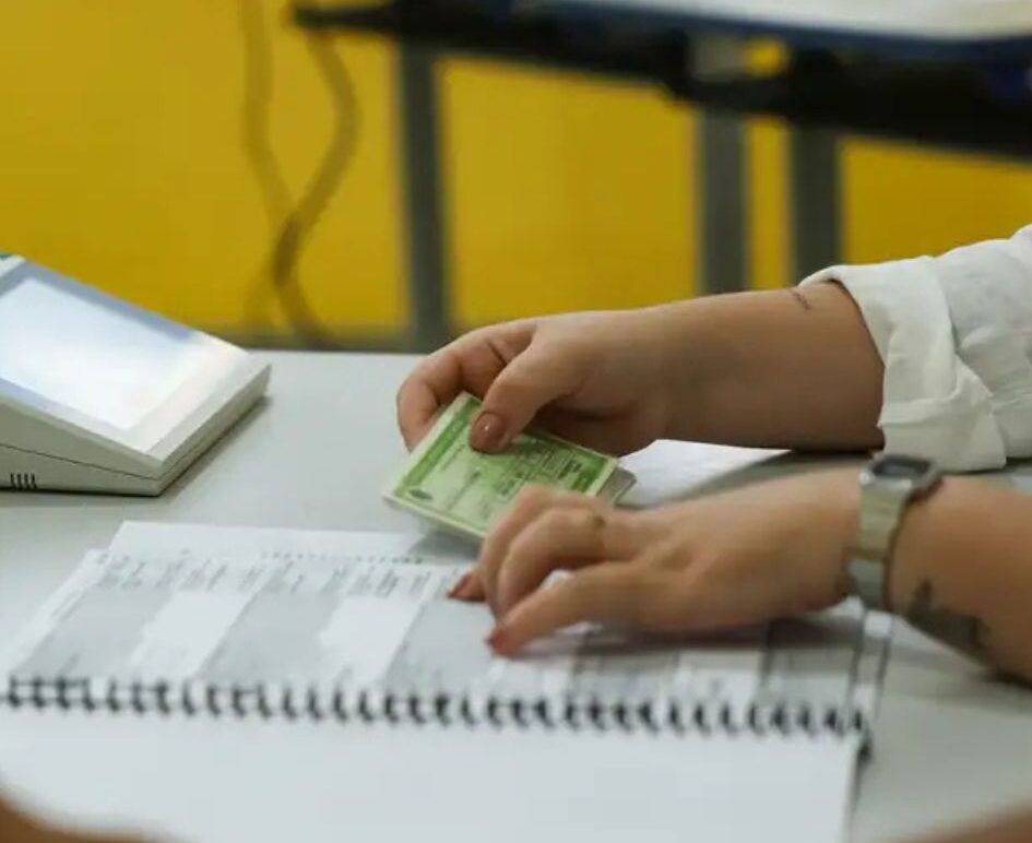 Fotografia em close das mãos de uma pessoa segurando um documento de identidade verde sobre um grande caderno de registros em espiral. À esquerda, levemente desfocado, aparece o terminal do mesário de uma urna eletrônica.
