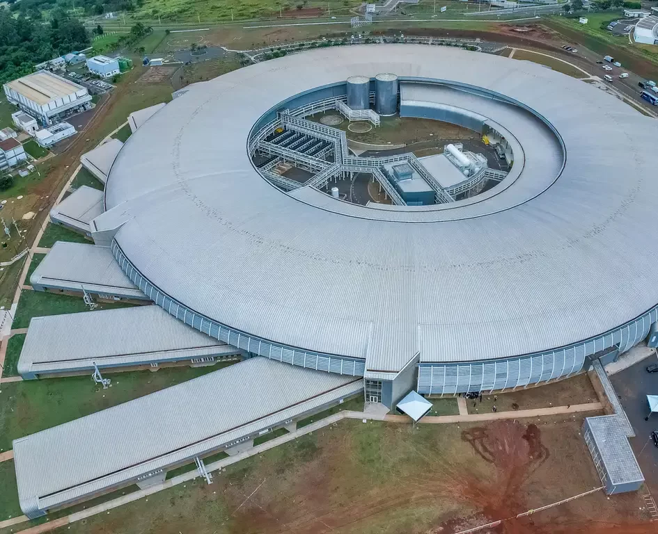 Vista aérea ampla do Laboratório Sirius em Campinas, exibindo toda a sua estrutura circular prateada e os prédios anexos de pesquisa, cercados por áreas verdes e infraestrutura rodoviária.