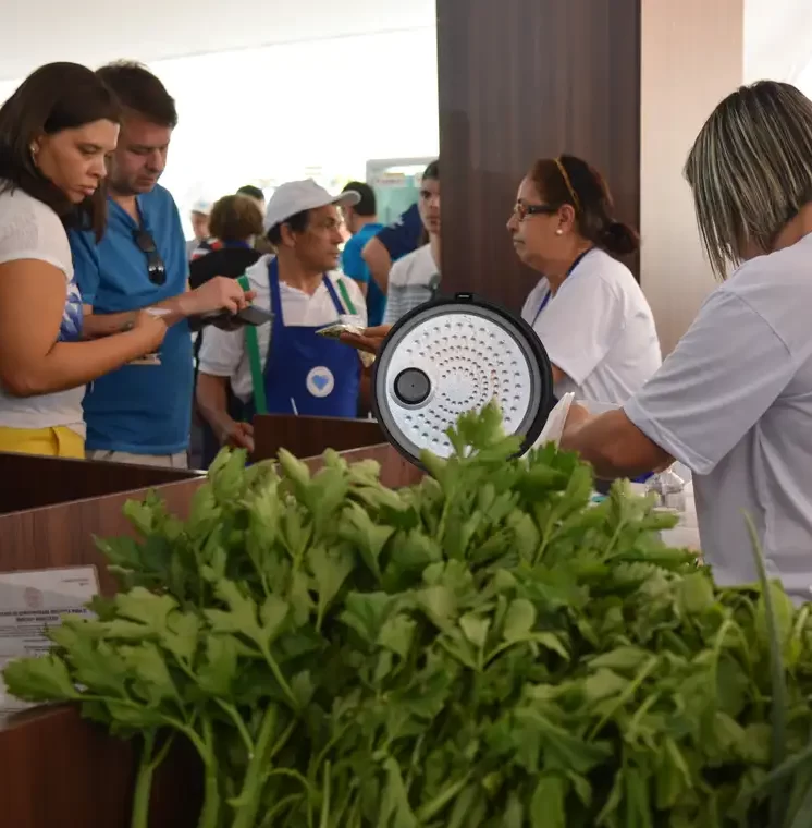 Fotografia de um maço de hortaliças verdes e frescas em primeiro plano, com pessoas interagindo e fazendo compras em barracas de uma feira livre ao fundo.