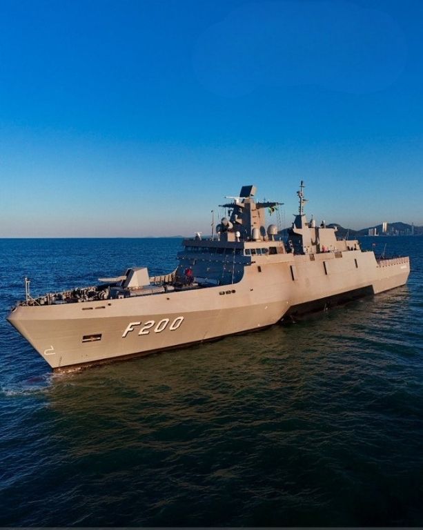 Fotografia de um grande navio de guerra cinza, com a inscrição "F200" em branco no casco, navegando em um mar de águas escuras sob um céu azul claro. O navio possui design moderno com linhas retas, um canhão na proa e uma torre central com radares. Ao fundo, à direita, há uma linha costeira distante com montanhas e silhuetas de prédios.