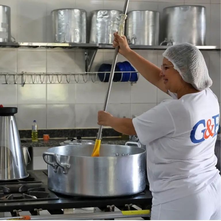 Uma mulher com touca de cabelo, óculos de segurança e uniforme branco com logo G&F está sorrindo enquanto mexe uma enorme panela de metal com uma espátula longa em uma cozinha industrial equipada.