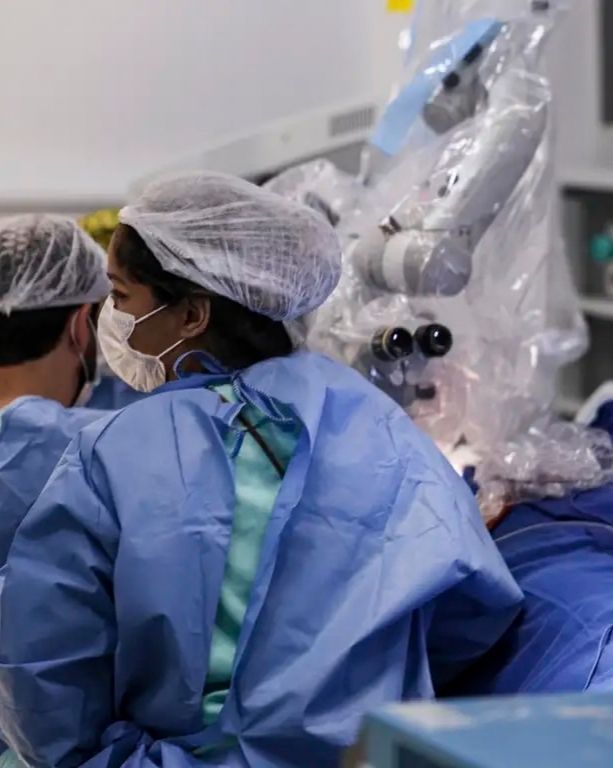 Uma fotografia de médio alcance de duas profissionais médicas em primeiro plano, de costas e perfil, vestindo trajes cirúrgicos azuis, toucas descartáveis e máscaras faciais brancas, focadas em um procedimento cirúrgico. À direita, um grande microscópio cirúrgico coberto por um protetor plástico estéril está visível, junto com equipamentos médicos de fundo e prateleiras de suprimentos.