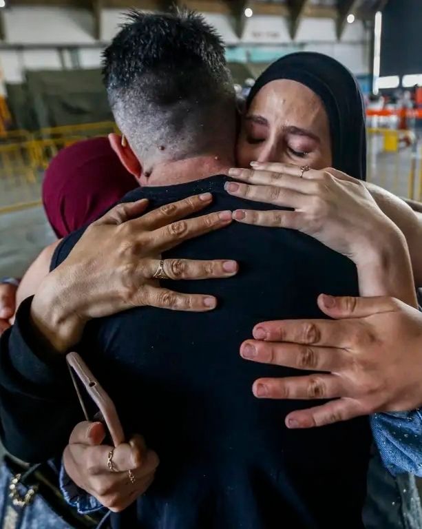 Fotografia de close-up de um abraço emocionado entre três pessoas em um terminal de transporte. No centro, as costas de um homem de camiseta preta e cabelo curto. Ele é abraçado por uma mulher (à direita) de hijab preto com olhos fechados e expressão de choro terno, cujas mãos com anéis estão sobre as costas dele. Outras mãos de diferentes tons de pele também se juntam ao abraço. O fundo desfocado mostra estruturas de teto de um terminal e luz natural.