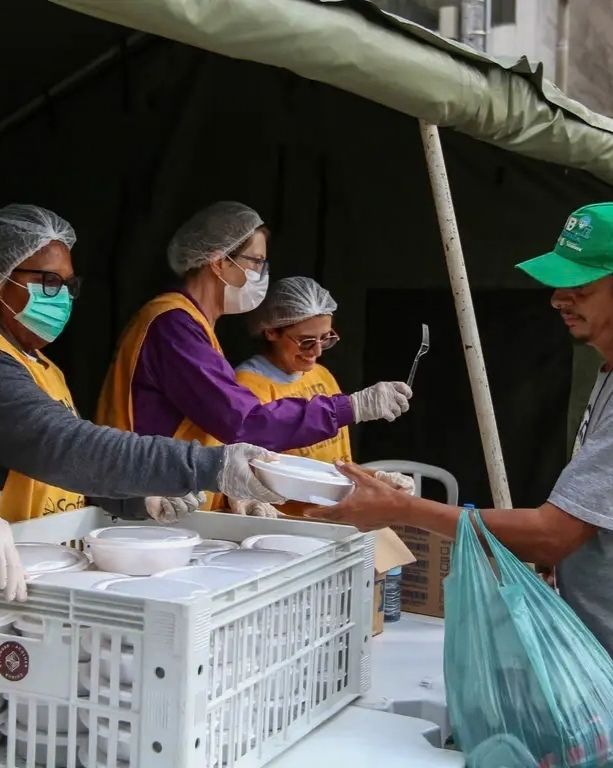 Fotografia de uma distribuição de alimentos sob uma tenda de lona verde. Três voluntárias com aventais amarelos, toucas descartáveis, máscaras faciais e luvas de proteção estão posicionadas atrás de duas caixas plásticas brancas cheias de marmitas embaçadas. Uma voluntária (extrema esquerda) está entregando uma marmita a um homem à direita, que usa um boné verde, camisa cinza e segura um saco plástico azul claro na mão esquerda. A voluntária central sorri para ele. O ambiente ao fundo é um acampamento ou área urbana desfocada.