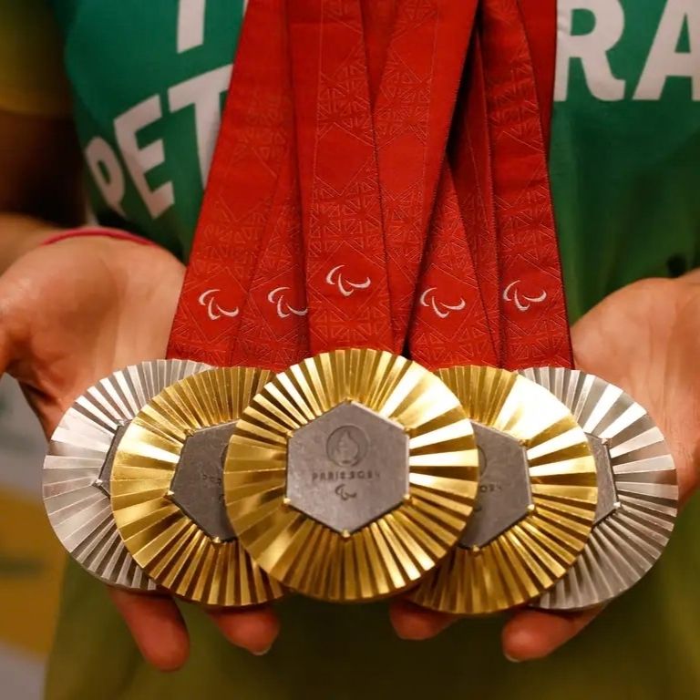Fotografia em close-up de duas mãos segurando cinco medalhas dos Jogos Paralímpicos de Paris 2024. No centro, destacam-se três medalhas de ouro, ladeadas por duas de prata. As medalhas possuem um centro hexagonal escuro com o logo de Paris 2024, cercado por sulcos radiantes. As fitas que as sustentam são vermelhas, largas, e trazem o símbolo branco dos Agitos Paralímpicos. O atleta veste uma camisa verde ao fundo, desfocada.