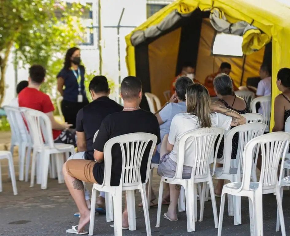 Um grupo de pessoas sentadas de costas para a câmera em cadeiras de plástico brancas em um pátio ao ar livre. Elas estão voltadas para uma palestrante que está em pé à esquerda. No fundo, à direita, há uma grande tenda de campanha amarela brilhante.