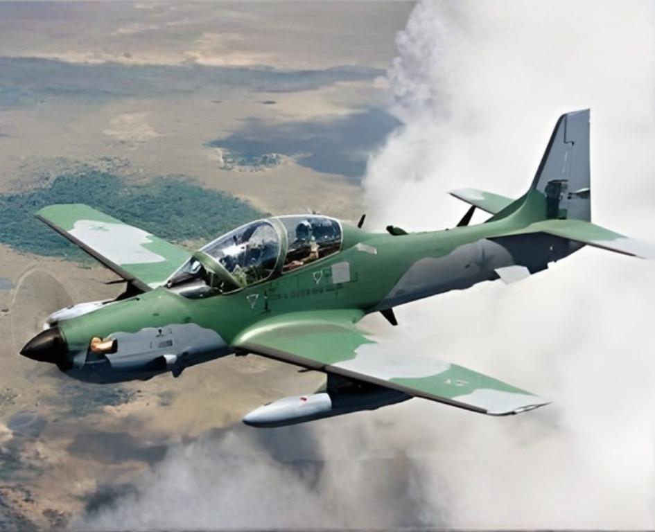 Fotografia em pleno voo de um avião militar turboélice A-29 Super Tucano. A aeronave possui camuflagem em tons de verde escuro e cinza claro. Há uma grande formação de nuvens brancas à direita e, ao fundo, vê-se a paisagem terrestre com planícies e vegetação.