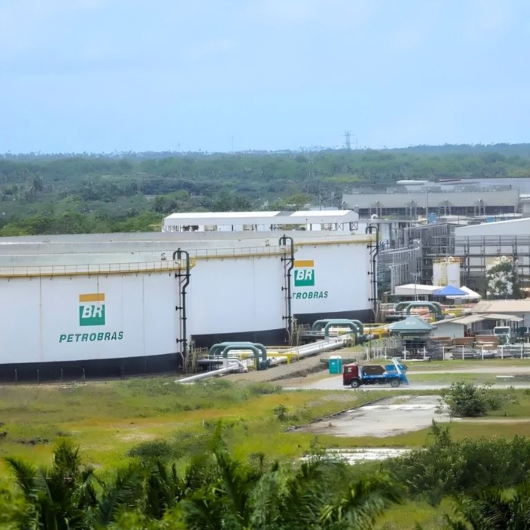 Uma vista aérea e panorâmica de uma grande instalação industrial da Petrobras no Brasil. Três grandes tanques cilíndricos brancos de armazenamento de combustível estão no centro, com os logotipos 'BR PETROBRAS' visíveis. Uma rede de tubulações e estruturas conecta os tanques e se estende para o fundo. Um caminhão de transporte pequeno se move por uma estrada de acesso. No fundo, há edifícios de processamento industrial e uma densa floresta tropical sob um céu nublado. A vegetação tropical rústica envolve o local.