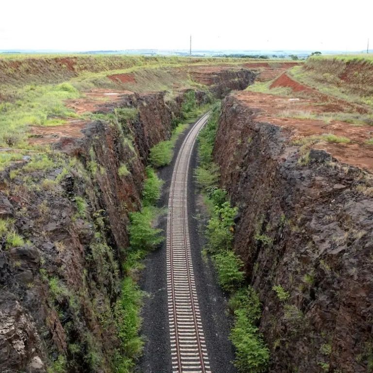 Uma vista aérea de uma única linha férrea com dormentes brancos, que se estende por um profundo corte em paredes de rocha escura e escarpada. O solo de terra vermelha é visível nas camadas superiores das encostas, e há vegetação rasteira verde crescendo ao longo das bases e paredes. A ferrovia se curva para a direita e desaparece em um desfiladeiro mais estreito ao fundo, com campos abertos e céu nublado no horizonte.