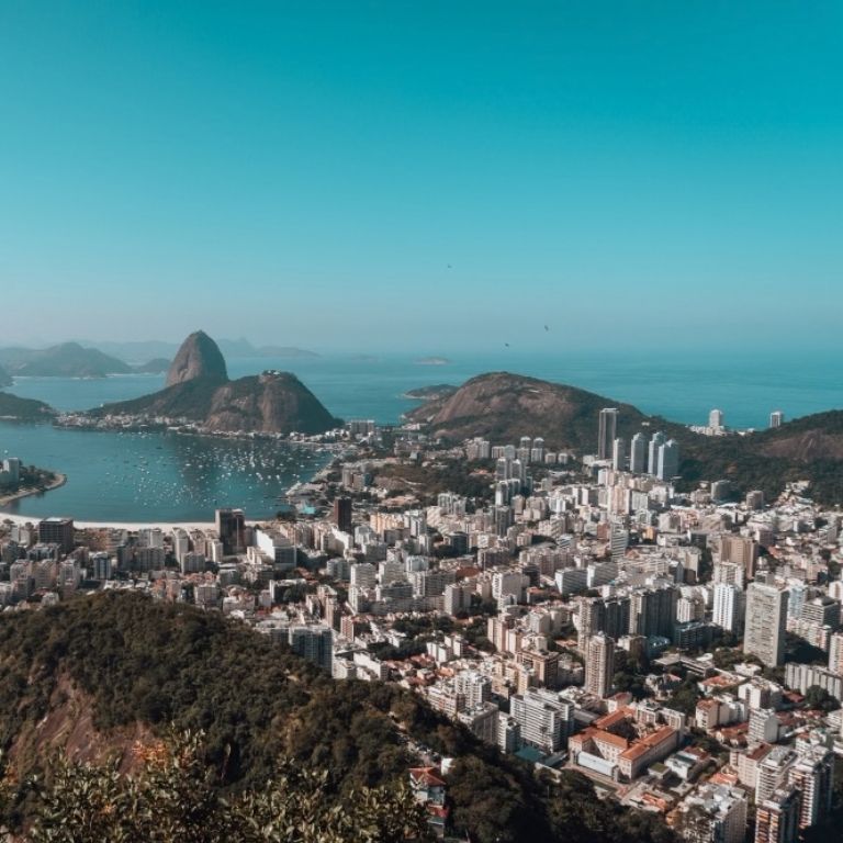 Vista aérea panorâmica de alta altitude da cidade do Rio de Janeiro, mostrando a Baía de Guanabara com muitos barcos ancorados, as icônicas montanhas do Pão de Açúcar e do Corcovado (com o Cristo Redentor visível no topo), e uma densa paisagem urbana sob um céu azul claro.