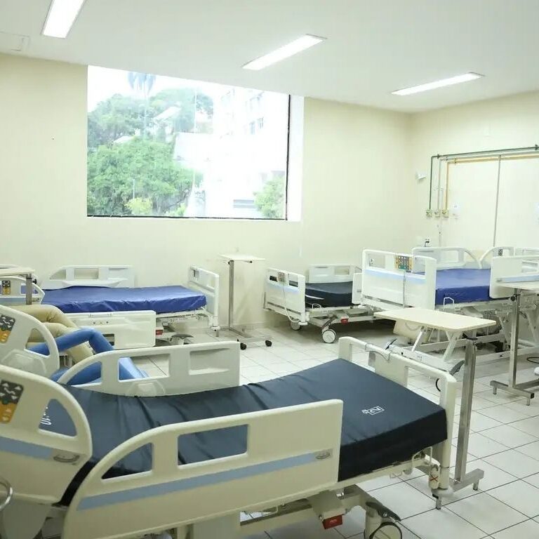 Quarto de hospital claro com várias camas hospitalares modernas vazias de colchão azul e uma janela ampla ao fundo com vista para árvores.