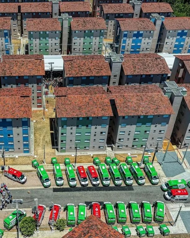 Fotografia aérea de ângulo alto capturando a escala de um grande conjunto habitacional de vários edifícios residenciais com telhados de cerâmica vermelha. Os edifícios têm seções de fachada colorida (azul e verde). Na rua abaixo, várias ambulâncias de emergência (principalmente verdes, com algumas vermelhas) estão estacionadas em paralelo. Uma pequena multidão de pessoas é visível.