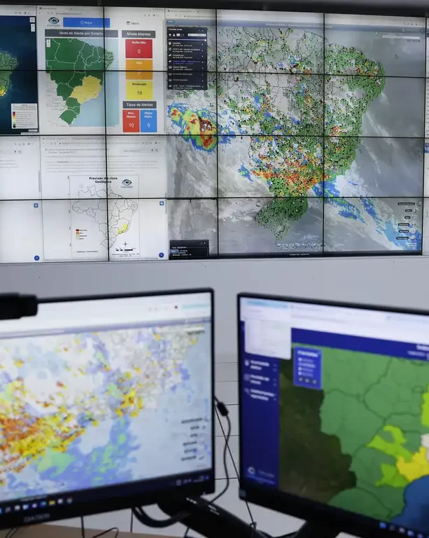 Centro de operações com múltiplos monitores exibindo mapas do Brasil com dados meteorológicos e geográficos coloridos, com um grande video wall ao fundo e dois monitores em primeiro plano.