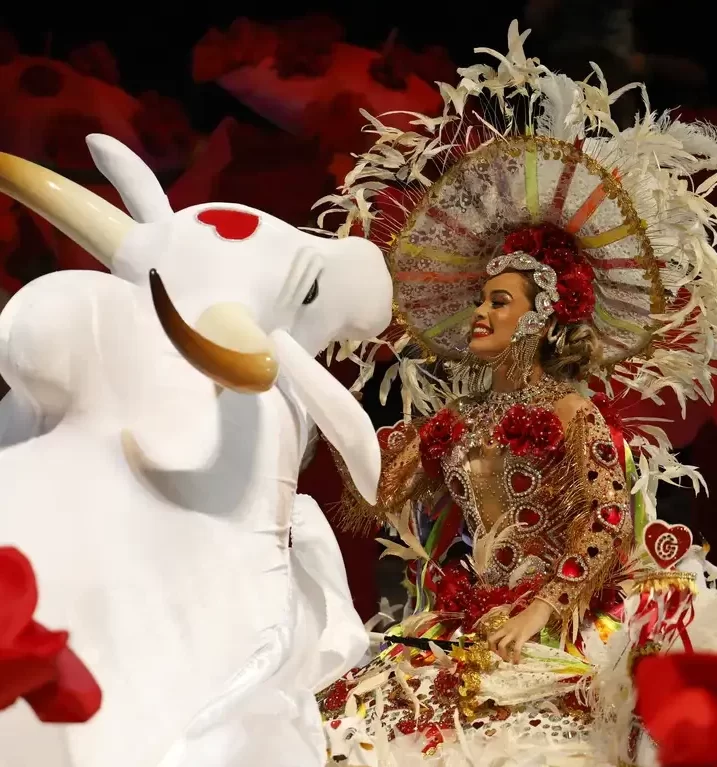 Integrante de festival folclórico sorridente, vestindo traje luxuoso com penas brancas e detalhes vermelhos, posicionada ao lado da figura do Boi Garantido (boi branco com um coração vermelho na testa).