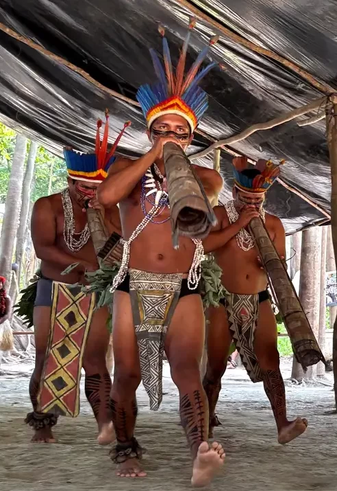 Três homens indígenas caminhando e tocando instrumentos de sopro longos e tradicionais. Eles usam cocares de penas coloridas, colares e tangas com grafismos geométricos sob uma estrutura de lona.