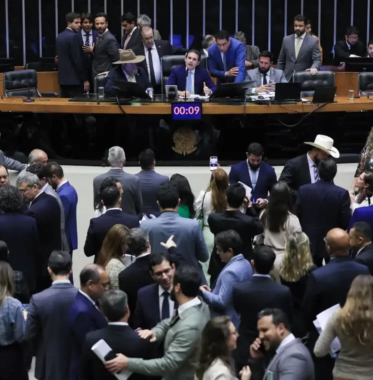 Fotografia de um plenário político lotado com dezenas de pessoas, a maioria homens vestindo ternos, conversando em pé na parte inferior. Na parte superior, há uma mesa de madeira elevada com o brasão da República do Brasil, onde alguns parlamentares estão sentados e em pé, discutindo. Um cronômetro digital vermelho no centro da bancada marca "00:09".