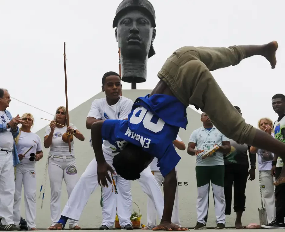 Jovem capoeirista realizando um movimento de "aú" (estrela) em frente ao busto do Monumento a Zumbi dos Palmares, com membros da roda de capoeira ao fundo.