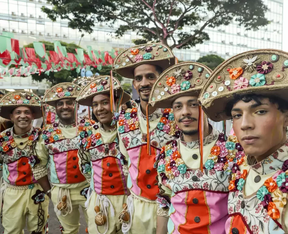 Cinco homens sorridentes vestindo trajes coloridos de festa popular e grandes chapéus de couro adornados com flores de fuxico. Ao fundo, bandeirinhas de festa junina e uma árvore.