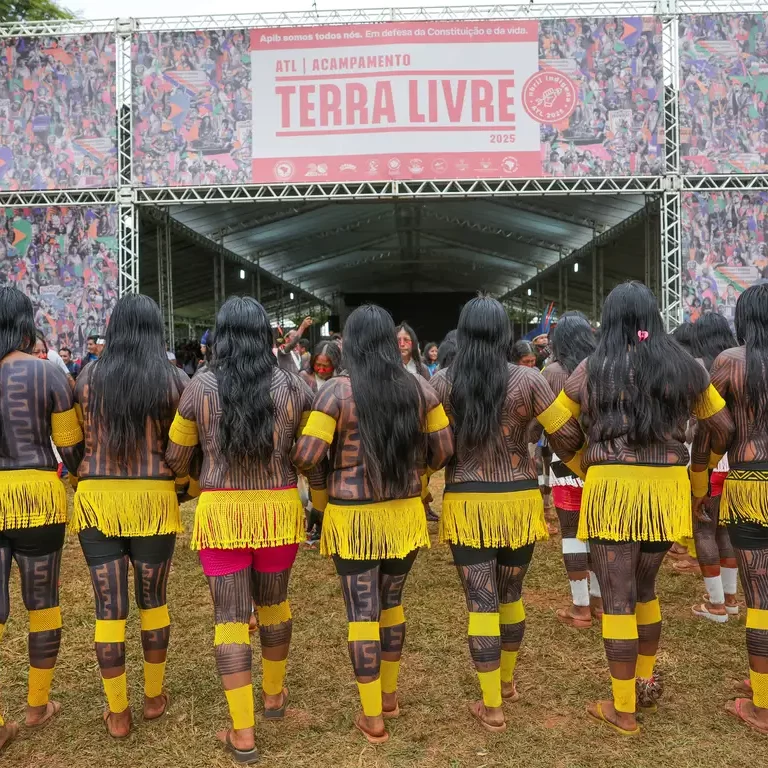 Mulheres indígenas de costas, com pinturas corporais tradicionais e saias de franjas amarelas, em frente a um grande palco com o banner do "ATL | Acampamento Terra Livre 2024".