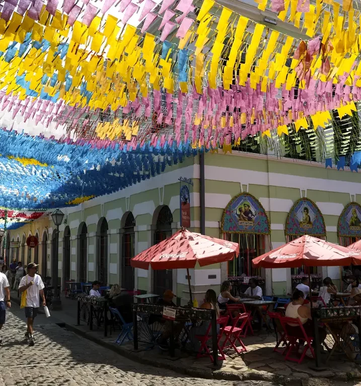 Rua decorada com centenas de bandeirinhas coloridas (amarelo, rosa, azul) suspensas sobre mesas de um restaurante ao ar livre em uma cidade histórica.