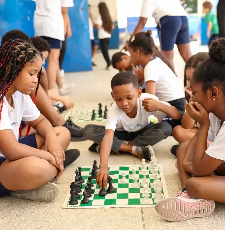 Crianças uniformizadas sentadas em círculo no chão, observando atentamente um menino que movimenta uma peça em um tabuleiro de xadrez verde e branco.