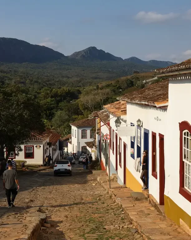 Rua de pedras em ladeira com casario colonial de fachadas brancas e molduras coloridas, com uma montanha imponente ao fundo sob céu azul.