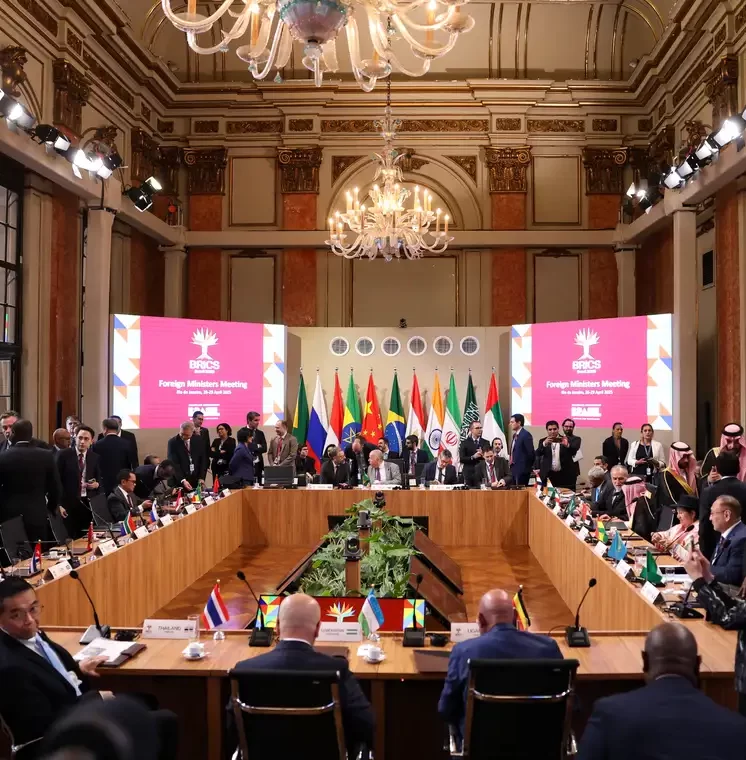 Fotografia ampla de um salão clássico e luxuoso com grandes lustres de cristal. No centro, dezenas de diplomatas e autoridades governamentais estão reunidos ao redor de uma grande mesa retangular de madeira. Ao fundo, há uma fileira com diversas bandeiras de países e dois telões cor-de-rosa com a logo em formato de árvore do BRICS e o texto em inglês "Foreign Ministers Meeting".
