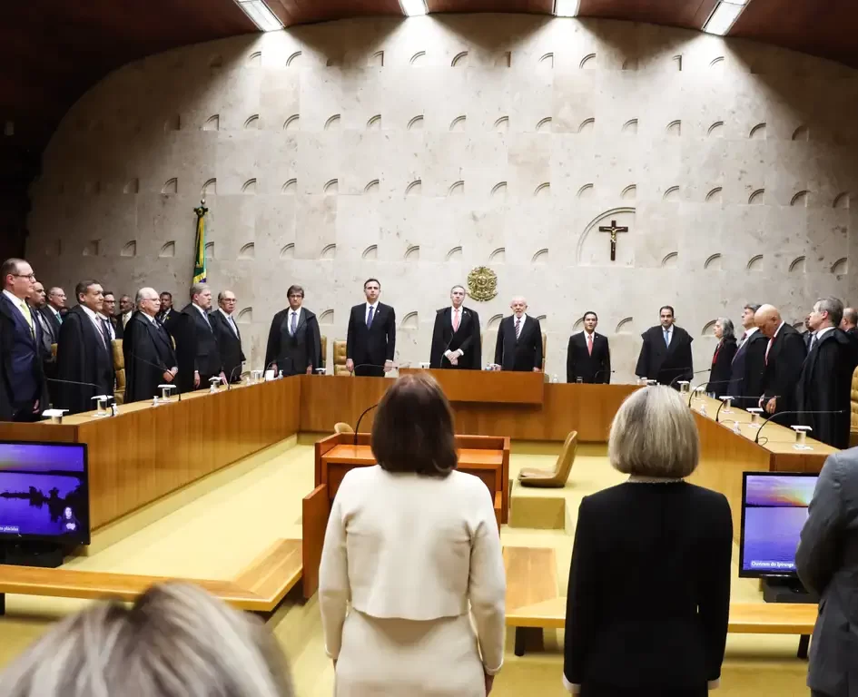 Visão de trás de um plenário do Supremo Tribunal Federal (STF) com Ministros em pé atrás da bancada principal de madeira vestindo togas pretas, com o brasão da República na parede de pedra ao fundo.