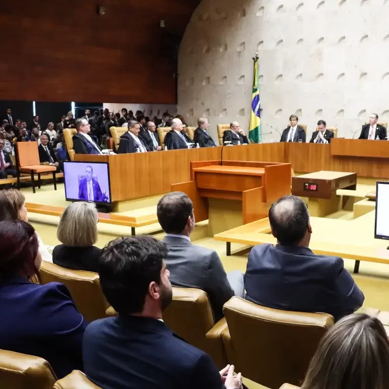 Fotografia do plenário do Supremo Tribunal Federal (STF). Ao fundo, ministros vestindo togas pretas estão sentados na bancada curva de madeira. À direita, há uma bandeira do Brasil. Em primeiro plano, pessoas na plateia assistem à sessão sentadas em poltronas de couro claro. À esquerda, uma tela exibe a transmissão em vídeo de um homem discursando.