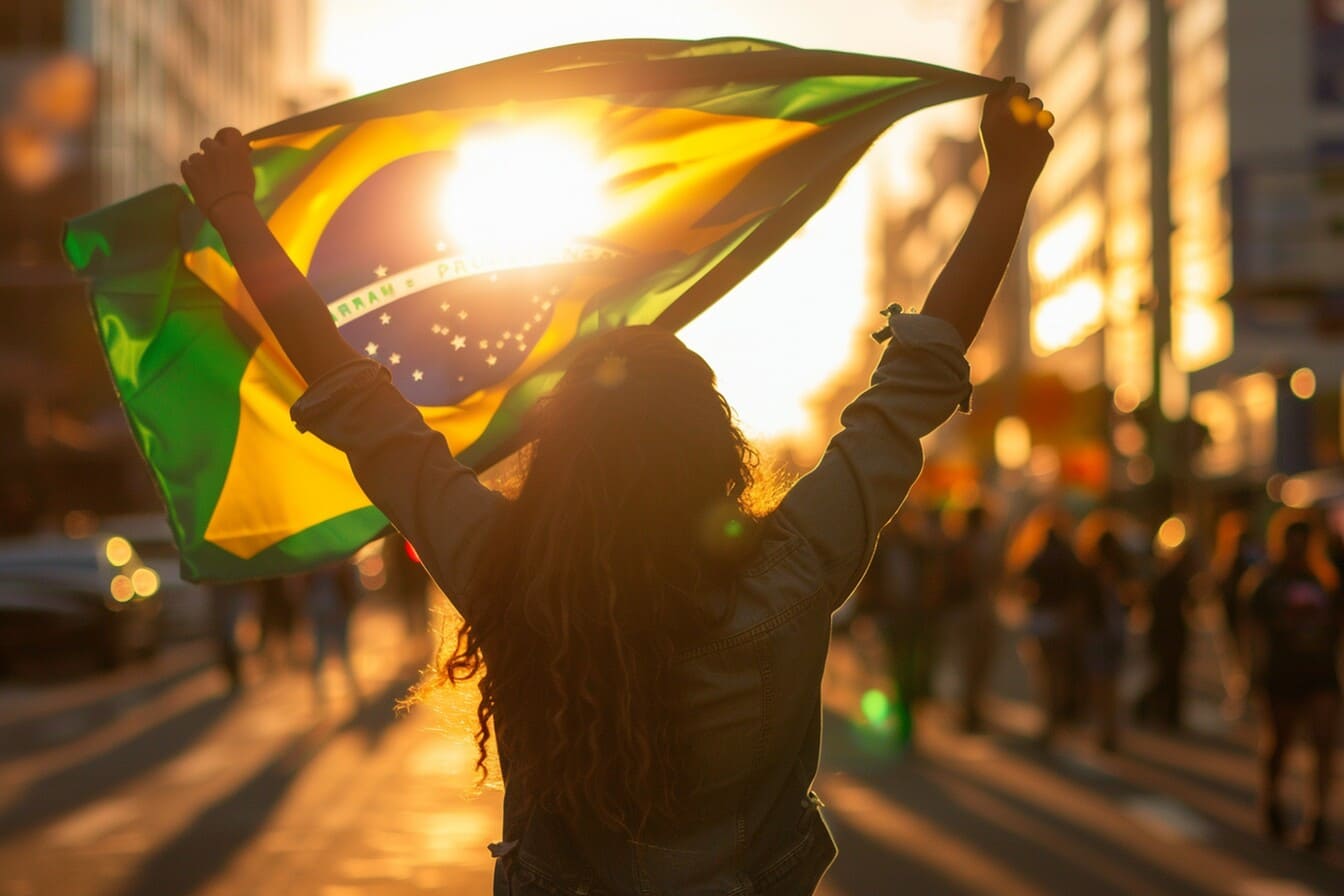 Mulher de costas segurando a bandeira do Brasil erguida com as duas mãos, silhuetada contra a luz forte do pôr do sol em uma avenida urbana movimentada.